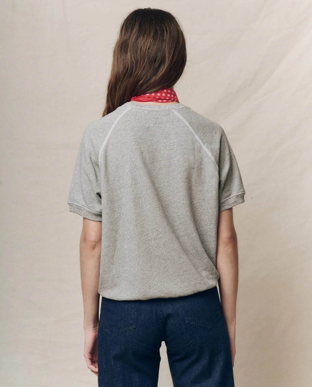 A person with long hair stands with their back to the camera, wearing The Great Inc.'s "The Short Sleeve Sweatshirt" in gray, blue jeans, and a red bandana around their neck against a neutral beige background.