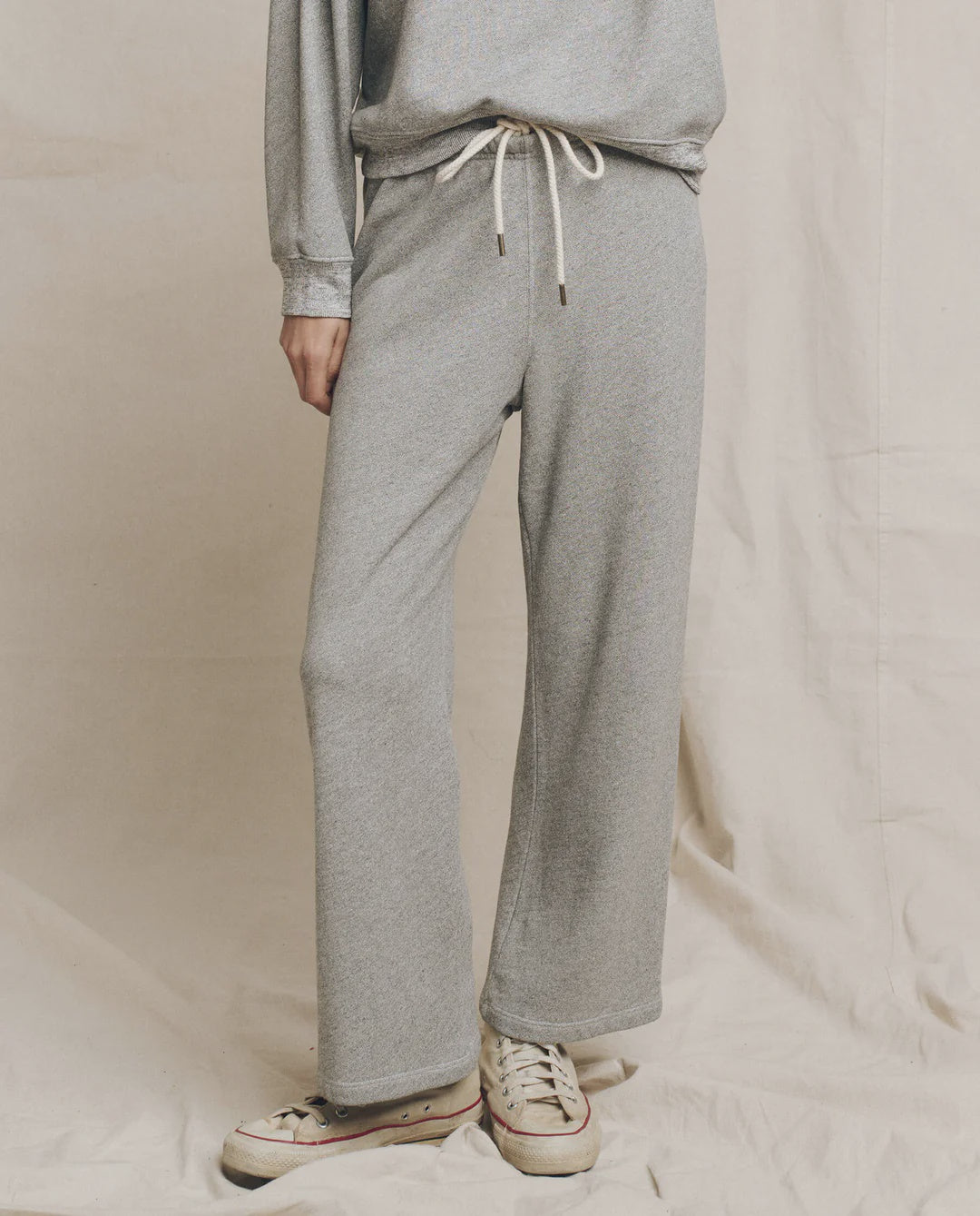 A person wears The Great Inc.'s The Lap Sweatpant and matching sweatshirt in light gray, paired with white sneakers, standing on a beige fabric backdrop. Only their body from the shoulders down is shown. This set is made in Los Angeles.