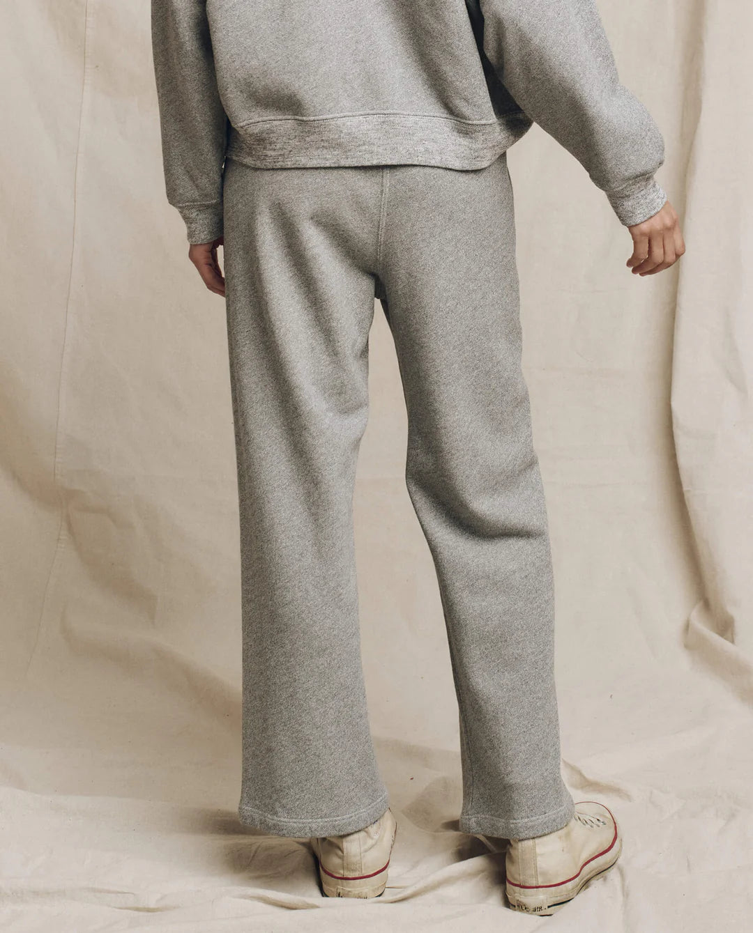 A person faces away, wearing The Great Inc.'s The Lap Sweatpant with a matching gray sweatshirt and off-white sneakers, set against a beige fabric backdrop. Every piece is handcrafted in Los Angeles.