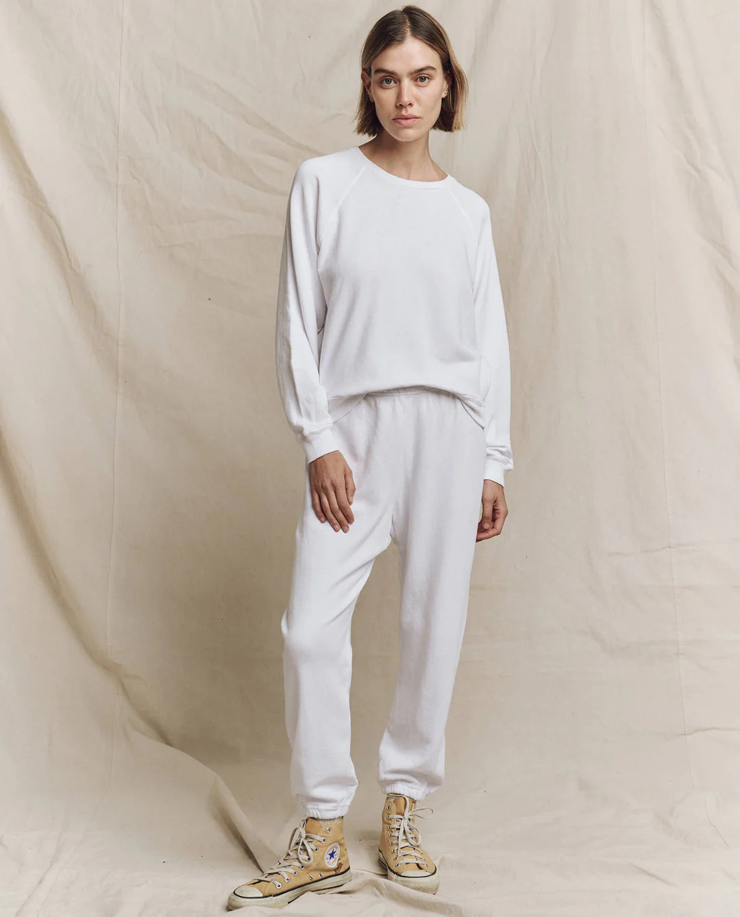 A person with short hair stands against a plain beige backdrop, wearing The College Sweatshirt from The Great Inc., featuring vintage graphics, and matching sweatpants. They sport light-colored high-top sneakers, creating a minimalistic, cozy, and relaxed vibe.