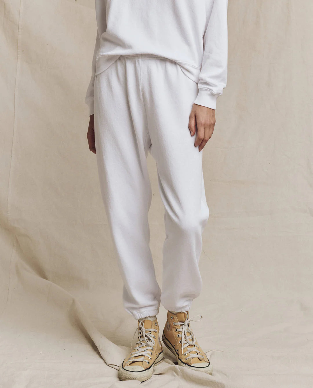 Dressed in a white long sleeve shirt and The Stadium Sweatpant by The Great Inc., the person stands against a neutral backdrop wearing beige high-tops with white laces, conveying a vintage athletic style. The image centers on their lower body.