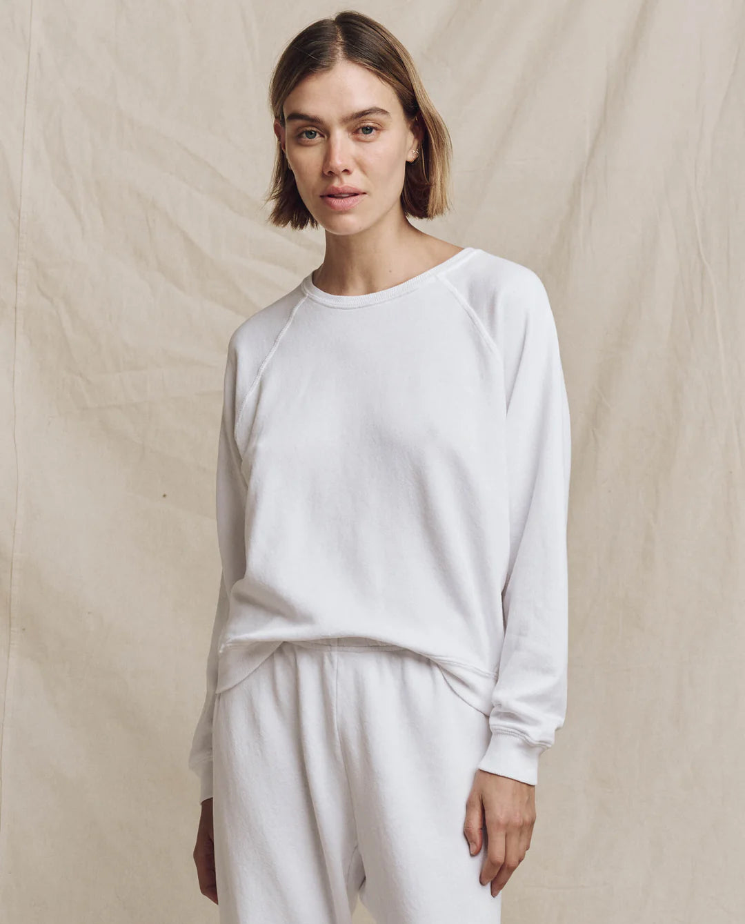 A person with short hair stands in front of a beige backdrop wearing The College Sweatshirt from The Great Inc., paired with pants, looking at the camera with a neutral expression.