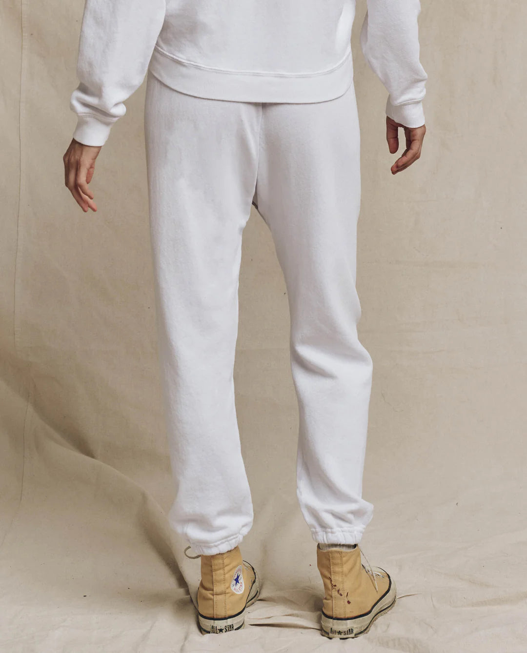 A person in a white sweatshirt and The Great Inc.'s Stadium Sweatpant stands with their back to the camera, wearing light brown high-top sneakers against a plain beige fabric background, capturing the simplicity of vintage athletic wear.