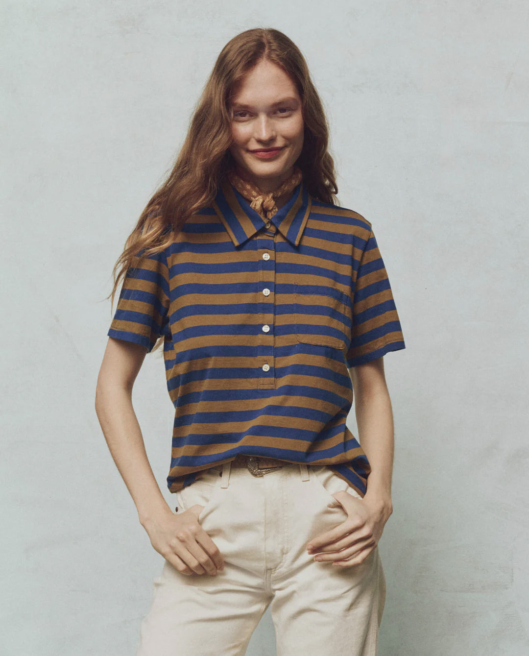 A person with long hair smiles with one hand on their hip, wearing The Lacrosse Polo by The Great Inc., featuring a brown and blue yarn dye stripe, made from 100% cotton, paired with light-colored pants against a light background.