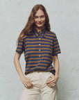 A person with long hair smiles with one hand on their hip, wearing The Lacrosse Polo by The Great Inc., featuring a brown and blue yarn dye stripe, made from 100% cotton, paired with light-colored pants against a light background.