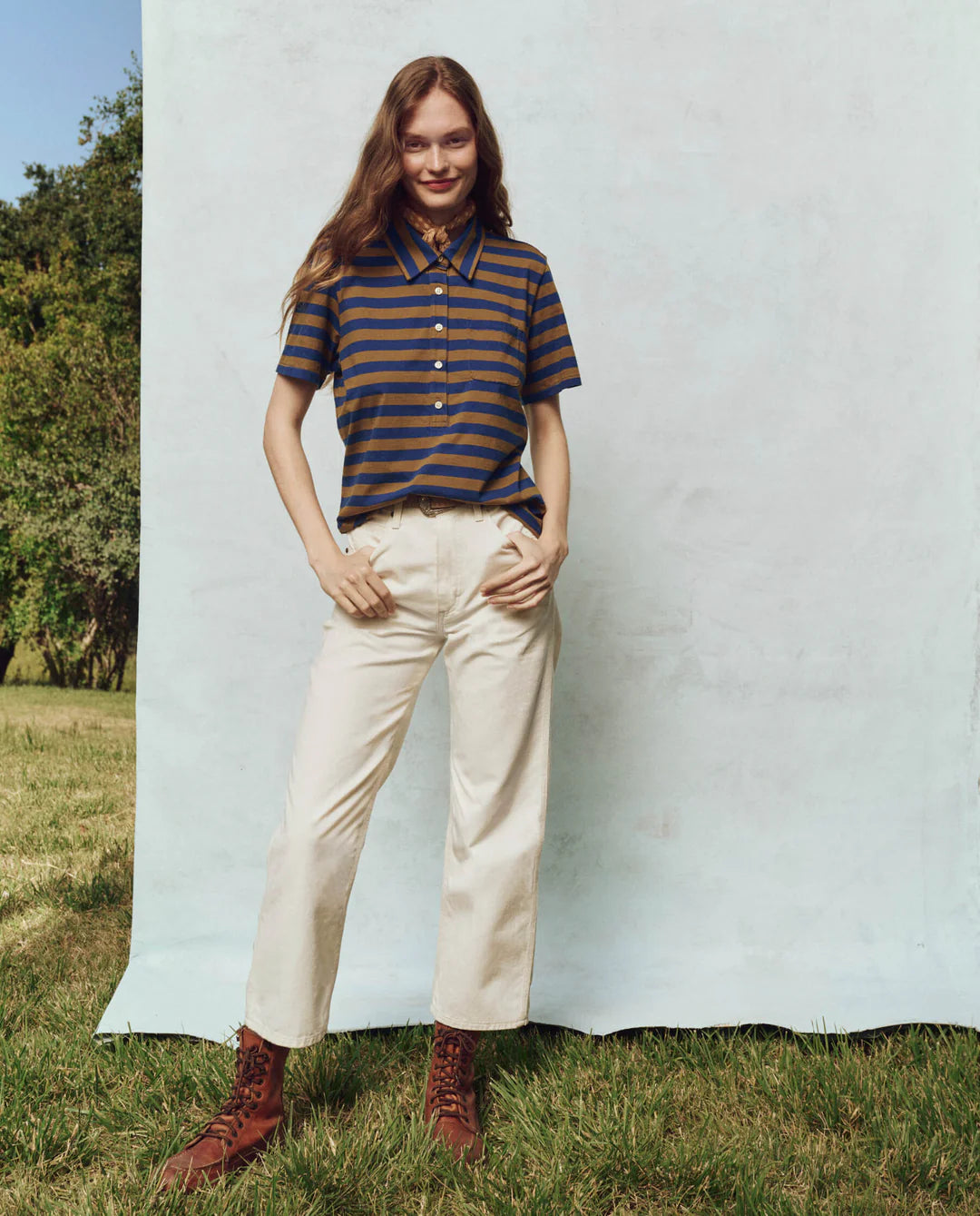 A person with long hair wears The Lacrosse Polo by The Great Inc., cream trousers, and brown boots, standing on grass. They pose with one hand in their pocket against a light backdrop, with trees forming a modern silhouette behind them.