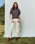 A person with long hair wears The Lacrosse Polo by The Great Inc., cream trousers, and brown boots, standing on grass. They pose with one hand in their pocket against a light backdrop, with trees forming a modern silhouette behind them.