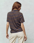 A person with long brown hair stands in The Great Inc.'s Lacrosse Polo, featuring blue and brown yarn-dye stripes, paired with light-colored pants. The backdrop is a plain, neutral tone.