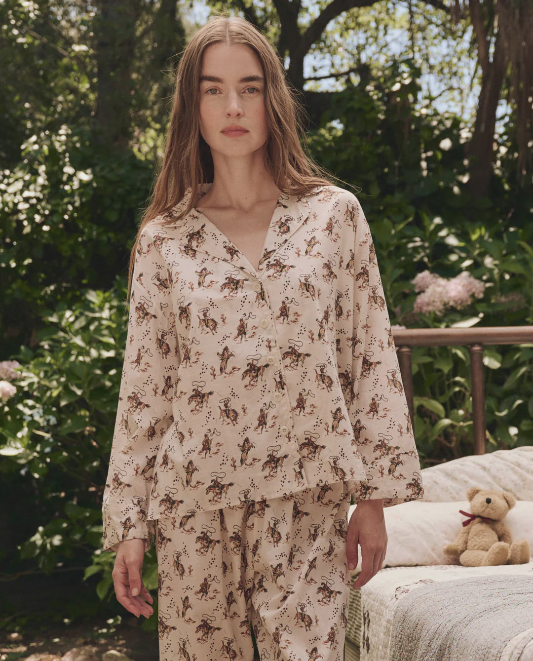 A woman with long brown hair stands outdoors in a lush garden wearing The Pajama Shirt by The Great Inc., a cream-colored top with a horse print and matching bottoms. A small teddy bear sits on a bench in the background, creating cozy vibes.