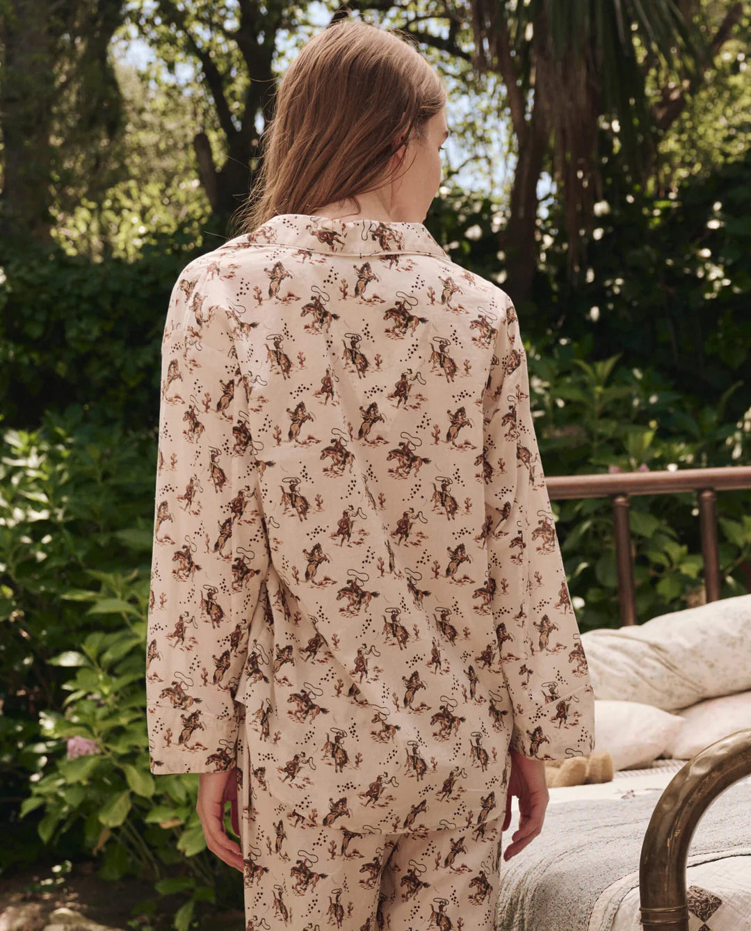 Seen from behind, a person with long hair stands outdoors in The Pajama Shirt by The Great Inc.—a beige top with a brown and green horse pattern. Lush foliage and a metal bed frame add to the vintage sleepwear vibe.