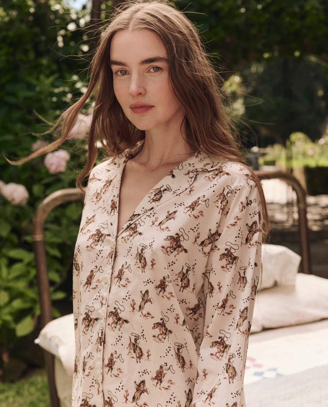 A woman with long brown hair stands outdoors, wearing The Pajama Shirt by The Great Inc., a light-colored blouse inspired by vintage pajama tops. Green foliage and part of a metal bed frame are visible in the background as she looks at the camera.