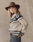 A person with long wavy blonde hair wears a brown cowboy hat, gray jeans, and The Snowflake Fairisle Pullover by The Great Inc., standing against a neutral fabric backdrop and gazing slightly to the side.
