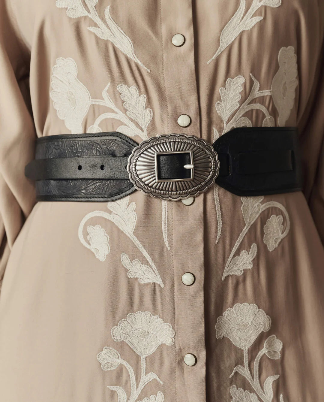 A close-up of a person wearing a beige shirt with white floral embroidery, accessorized with The Great Inc. Tooled Waist Belt featuring an ornate, oval silver buckle.