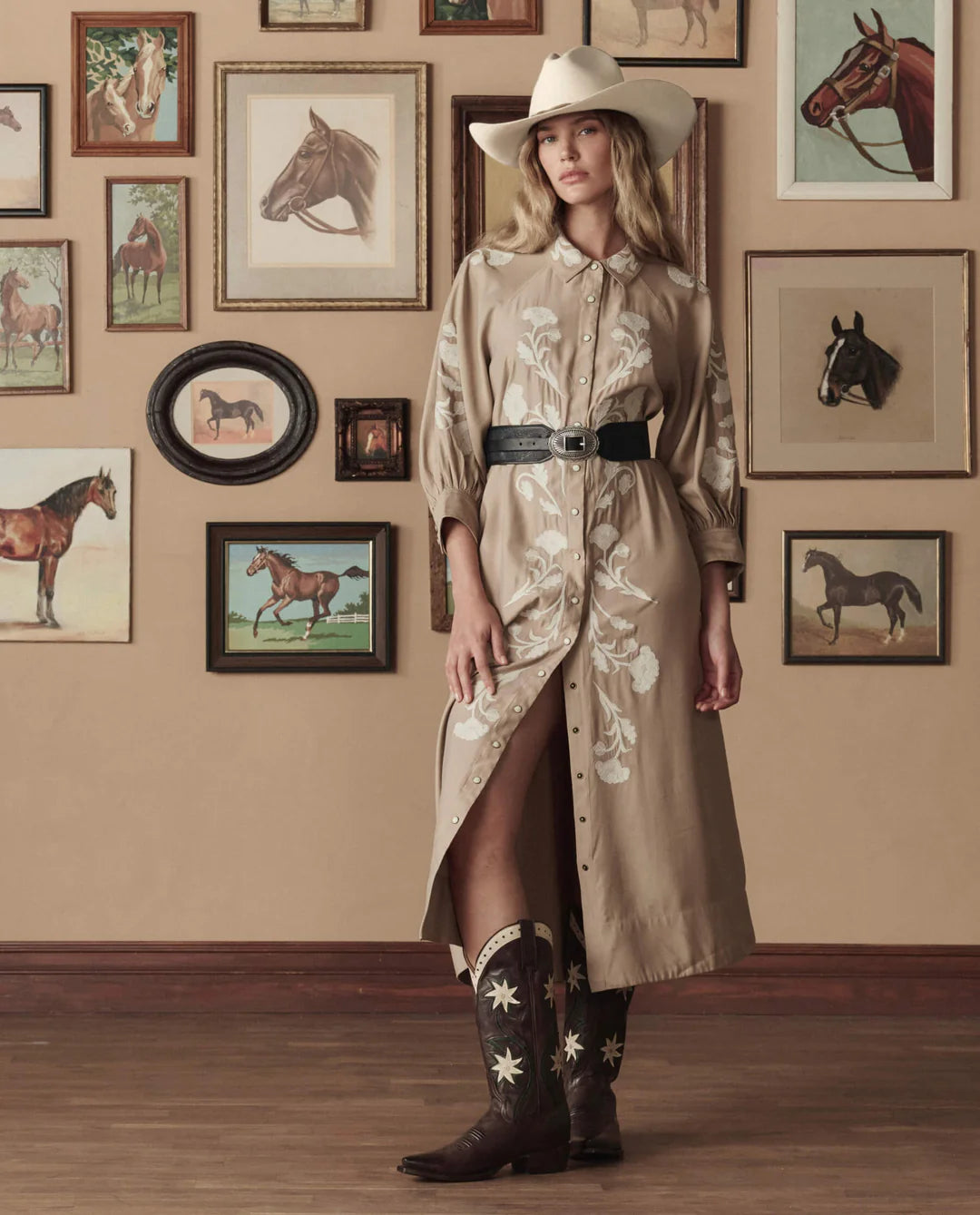 A woman in a beige cowboy hat, embroidered tan dress, wide black belt, and The Great Inc. Leather Floral Cowboy Boot stands indoors by a wall of framed horse art. Wood floors and decor evoke a Western-inspired mood.