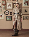 A woman in a beige cowboy hat, embroidered tan dress, wide black belt, and The Great Inc. Leather Floral Cowboy Boot stands indoors by a wall of framed horse art. Wood floors and decor evoke a Western-inspired mood.