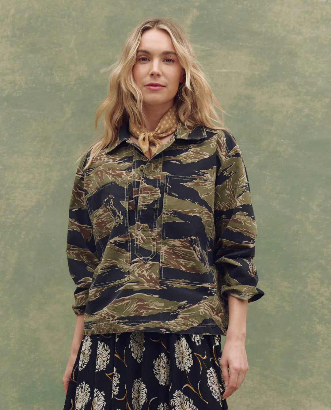 A woman with wavy blonde hair wears The Great Inc. The Railway Shirt over a floral skirt and a tan scarf, standing against a green, textured background.