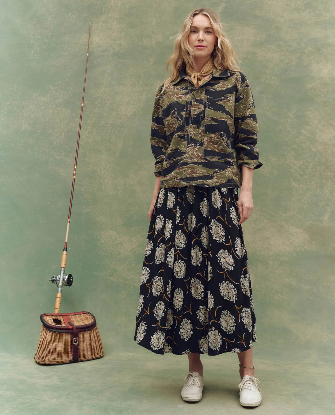 A woman stands before a mottled green backdrop in The Railway Shirt by The Great Inc., paired with a long navy floral skirt, white shoes, and a patterned scarf. A fishing rod and basket nearby add vintage workwear charm.
