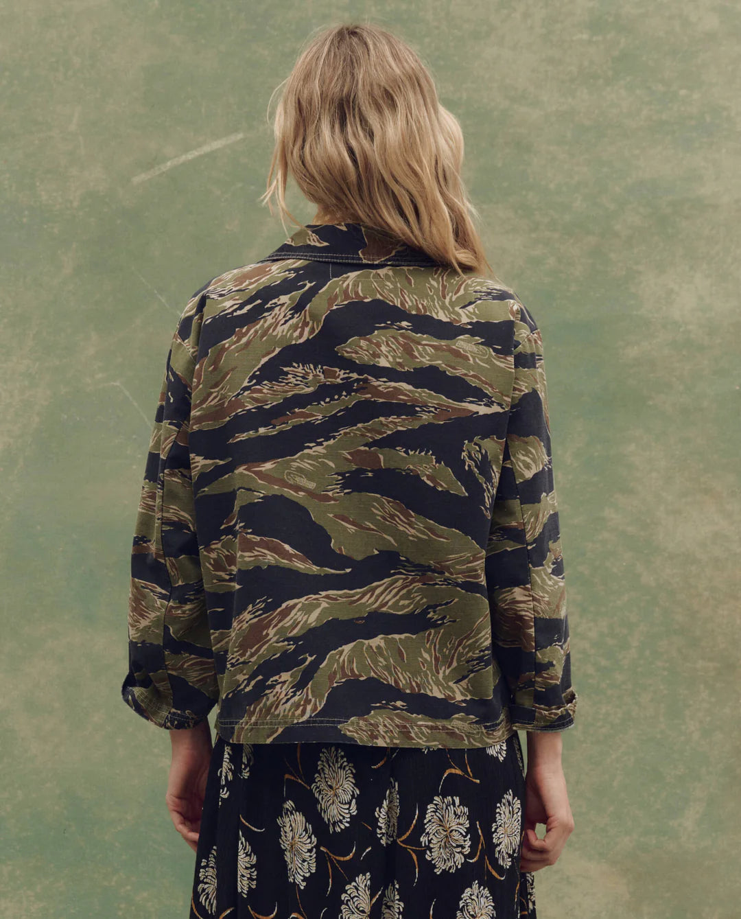A person with long, wavy blonde hair stands facing away, wearing The Railway Shirt by The Great Inc.—a green and brown tiger-striped jacket—and a black skirt with a white floral print against a muted green background.
