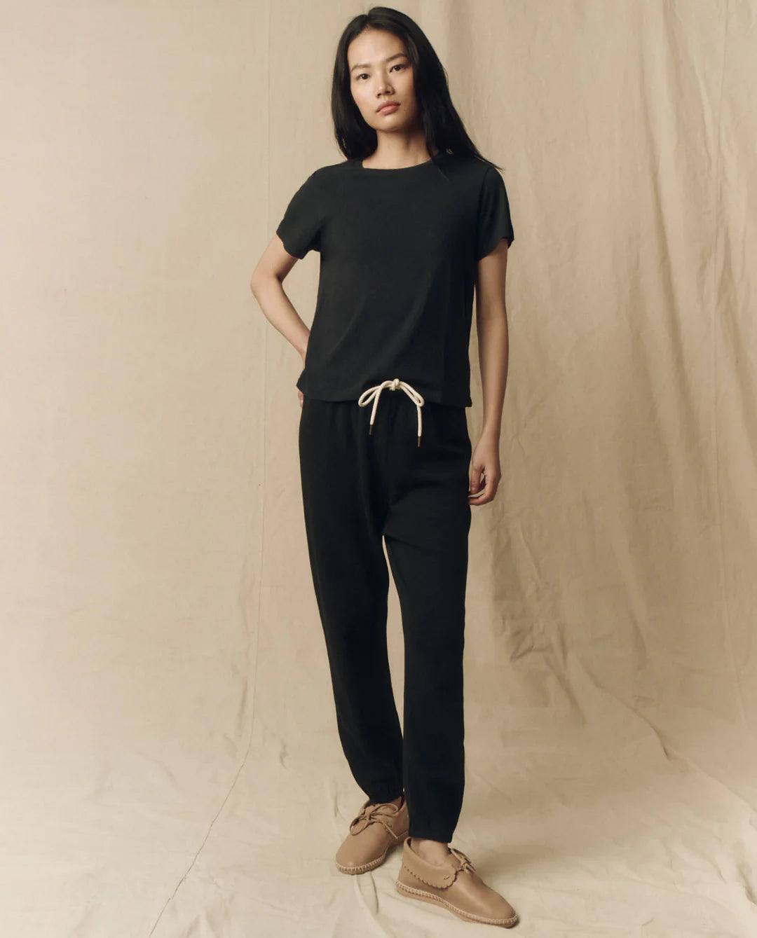 A woman with long straight hair and a neutral expression wears The Little Tee by The Great Inc., black drawstring pants with a cream tie, and tan moccasin-style shoes, standing against a beige fabric backdrop. This look is made in L.A.