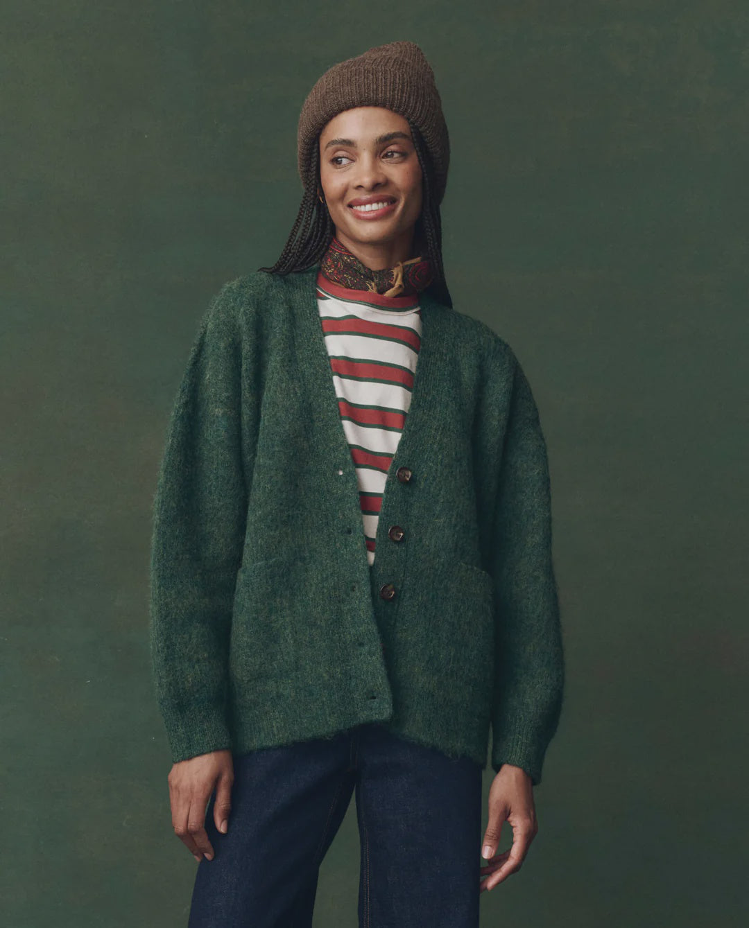 A person wearing The Great Inc. Brushed Melange Cardigan, paired with a striped shirt, dark jeans, a brown knit beanie, and a patterned scarf stands against a green background, smiling and looking slightly to the side.