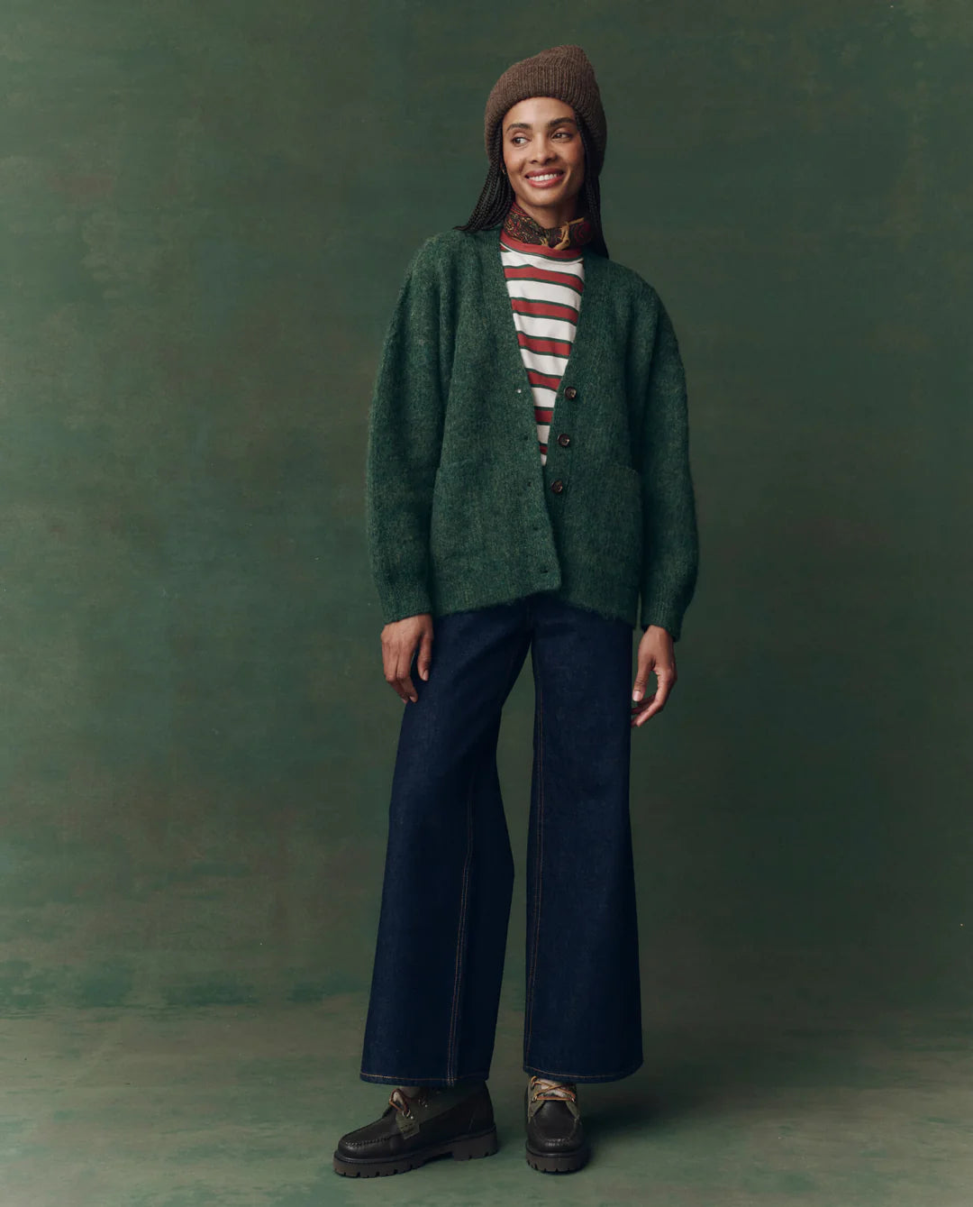 A person stands smiling against a green background, wearing The Brushed Melange Cardigan by The Great Inc. over a red-and-white striped shirt, dark wide-leg jeans, brown shoes, a patterned scarf, and a knitted brown beanie.