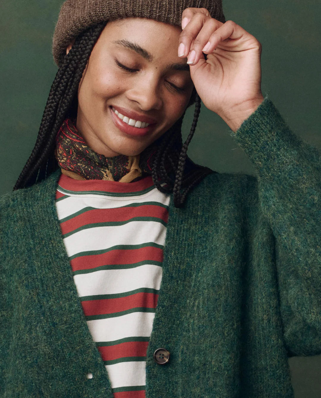 A woman with braids smiles with eyes closed, wearing The Brushed Melange Cardigan by The Great Inc., a brown knit hat, a striped red and white shirt, and a patterned neck scarf against a green background.