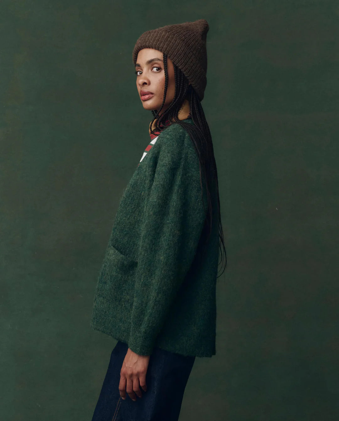 A person with long braids wears The Brushed Melange Cardigan by The Great Inc. in dark green, paired with a brown knitted beanie and dark pants, standing sideways against a dark green background.