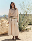 A woman poses in a desert landscape near agave plants and rocks, wearing The Great Inc.’s The Crochet Pullover, paired with a long cream skirt, a decorated belt, and brown boots under a clear blue sky.