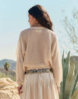 A woman with long, dark hair stands outdoors among desert plants and hills, her back to the camera. She's wearing The Crochet Pullover by The Great Inc. over a flowy skirt and decorative belt.