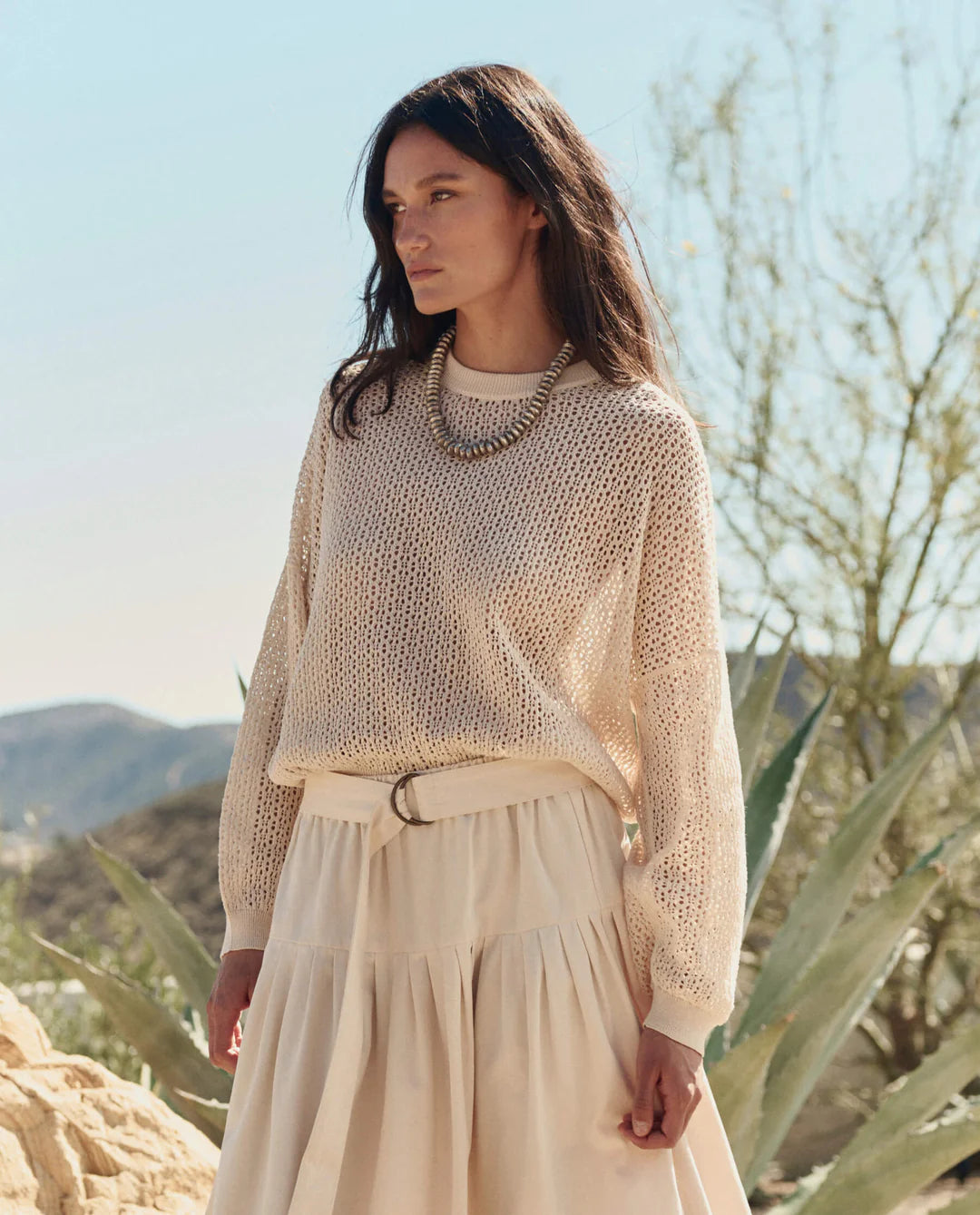 A woman wearing The Crochet Pullover by The Great Inc. stands outdoors amid desert plants and mountains, paired with a flowy beige skirt and a chunky necklace.
