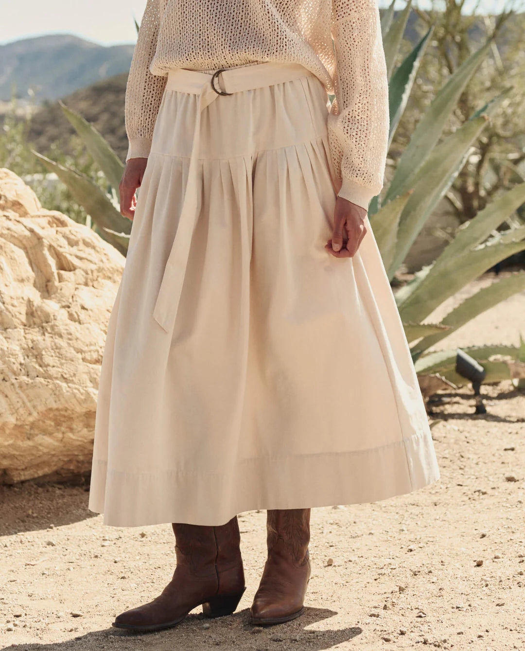Wearing The Highland Skirt by The Great Inc., a cream cotton canvas piece with a vintage feel, this person pairs it with a textured sweater and brown cowboy boots, standing on sandy ground among desert plants and rocks.