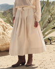Wearing The Highland Skirt by The Great Inc., a cream cotton canvas piece with a vintage feel, this person pairs it with a textured sweater and brown cowboy boots, standing on sandy ground among desert plants and rocks.