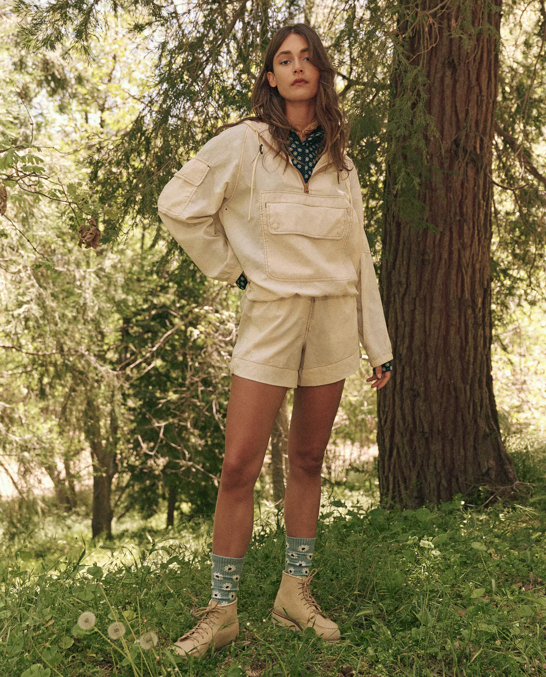 Amongst the forest's lush foliage, a person confidently wears The Trailhead Pullover by The Great Inc., along with durable shorts. Patterned socks and matching shoes complete the ensemble as sunlight highlights their technical clothing, with one hand resting casually on their hip.