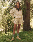 Amongst the forest's lush foliage, a person confidently wears The Trailhead Pullover by The Great Inc., along with durable shorts. Patterned socks and matching shoes complete the ensemble as sunlight highlights their technical clothing, with one hand resting casually on their hip.