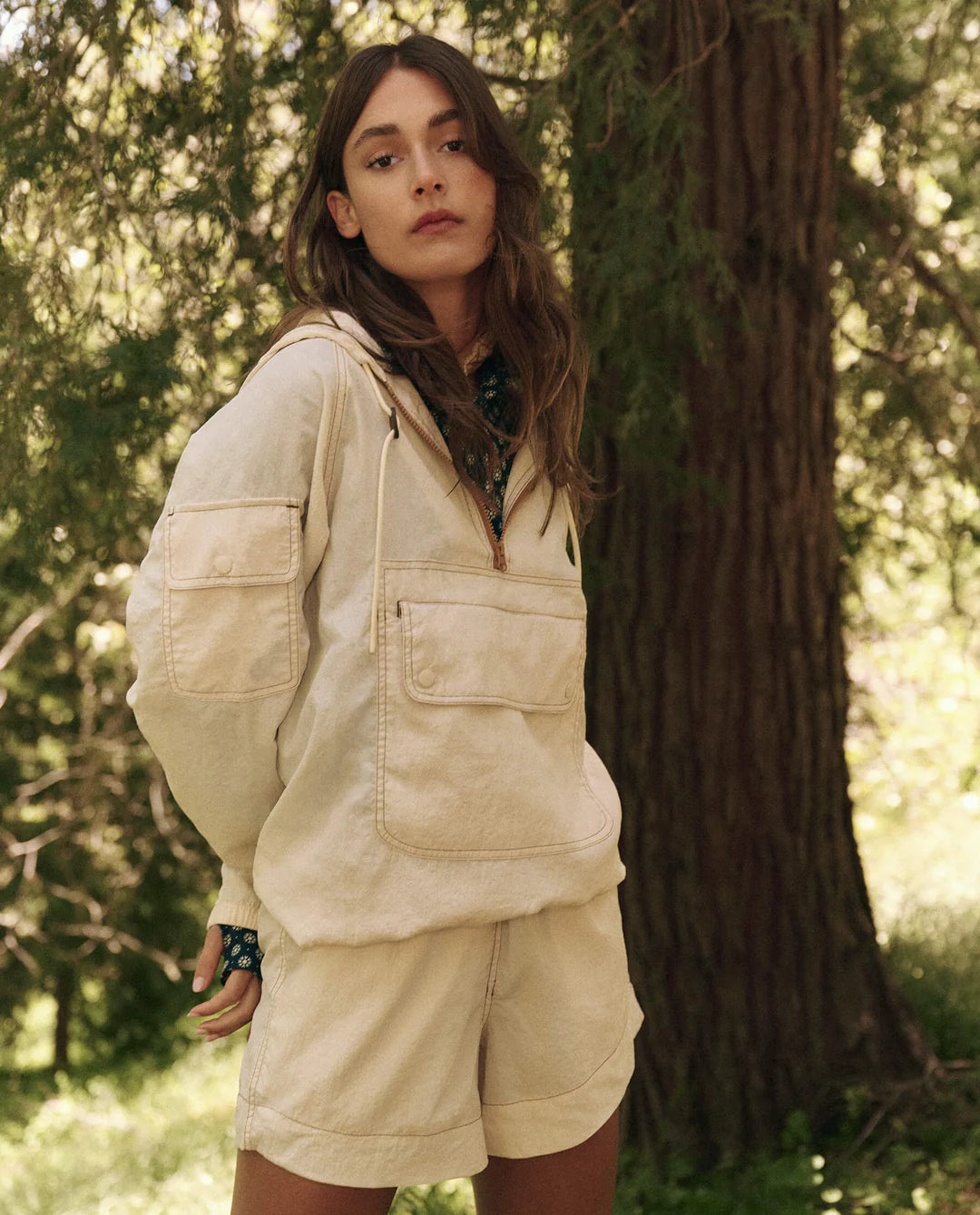 A person in The Great Inc.&#39;s light beige Trailhead Pullover, crafted from durable fabric, stands outdoors near trees. With long brown hair and a neutral expression, hands partly in pockets, the scene evokes a serene, natural setting.