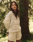 A person outdoors in The Rover Short by The Great Inc. pairs a pocketed jacket with the light-colored, water-repellant shorts amid lush greenery and tall trees, offering a serene forest setting ideal for showcasing this technical clothing.