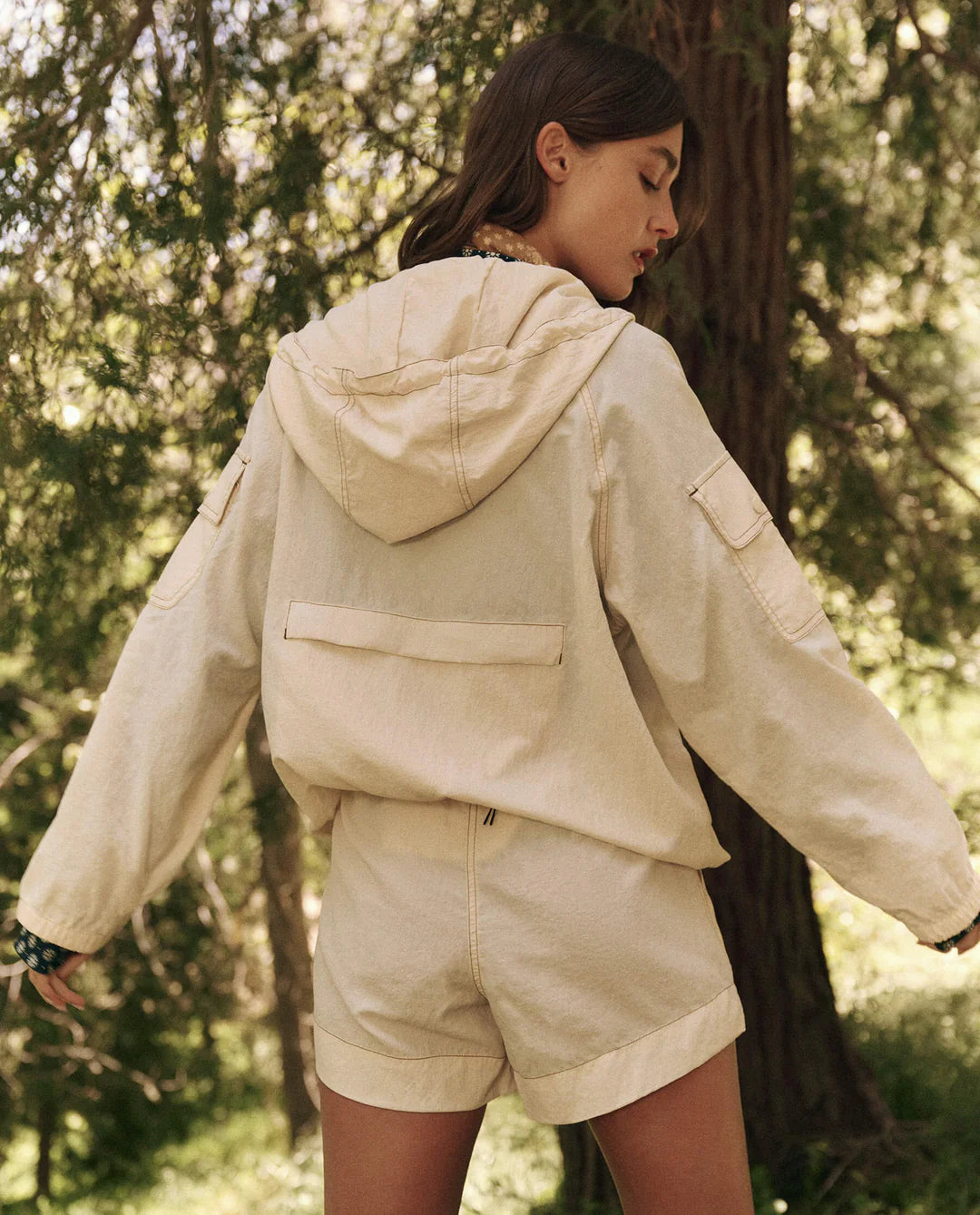 A woman stands in a forest, wearing The Trailhead Pullover by The Great Inc., a beige hooded jacket paired with matching shorts made from durable fabric. She faces away, her head turned slightly to the side as sunlight filters through the trees, casting dappled shadows around her.