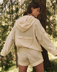 A woman stands in a forest, wearing The Trailhead Pullover by The Great Inc., a beige hooded jacket paired with matching shorts made from durable fabric. She faces away, her head turned slightly to the side as sunlight filters through the trees, casting dappled shadows around her.