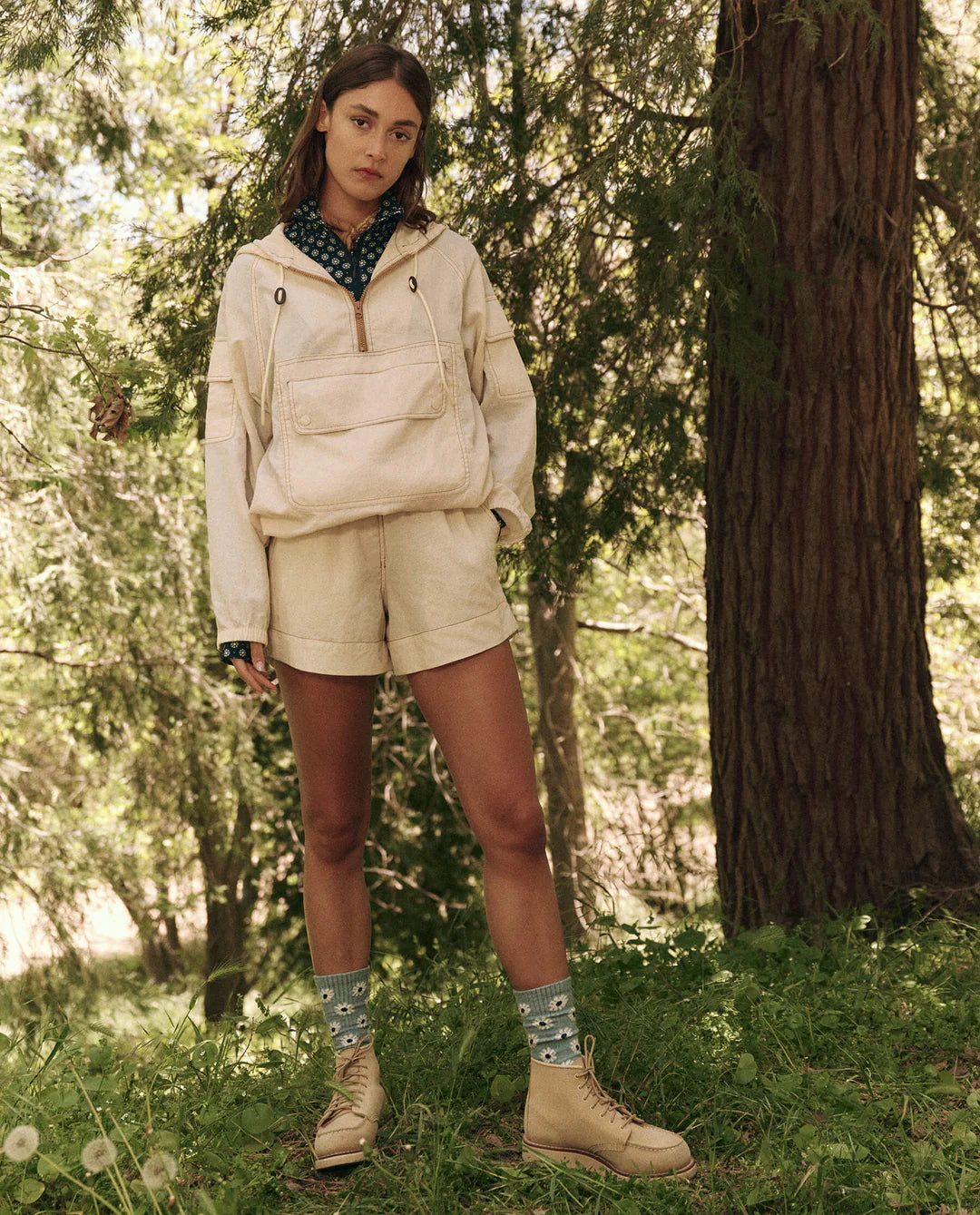 In a wooded area, an individual confidently wears The Great Inc.'s light beige jacket with durable fabric and The Rover Short. Blue patterned socks peek above tan boots as they stand on grass, surrounded by trees, looking directly at the camera.