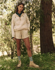 In a wooded area, an individual confidently wears The Great Inc.'s light beige jacket with durable fabric and The Rover Short. Blue patterned socks peek above tan boots as they stand on grass, surrounded by trees, looking directly at the camera.