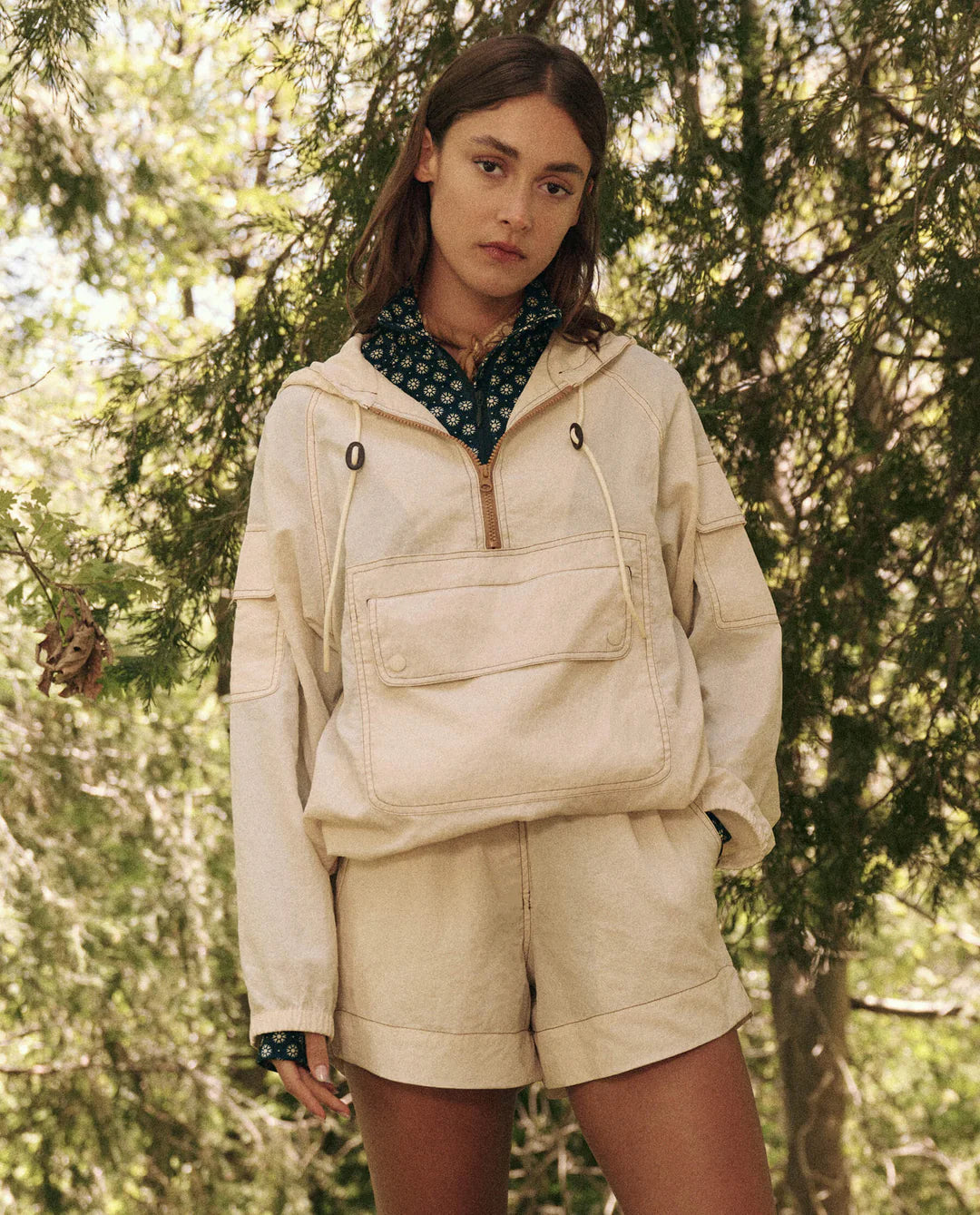 A person wears The Great Inc.'s Trailhead Pullover, light-colored and made of durable fabric, standing outdoors with long brown hair and a neutral expression. They're surrounded by lush green trees and foliage in a natural setting.