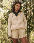 A person wears The Great Inc.'s Trailhead Pullover, light-colored and made of durable fabric, standing outdoors with long brown hair and a neutral expression. They're surrounded by lush green trees and foliage in a natural setting.