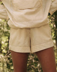 Wearing "The Rover Short," a person stands outdoors in a forested area, showcasing this durable beige hoodie and shorts combo by The Great Inc. Amidst blurred greenery, the focus remains on the technical clothing.