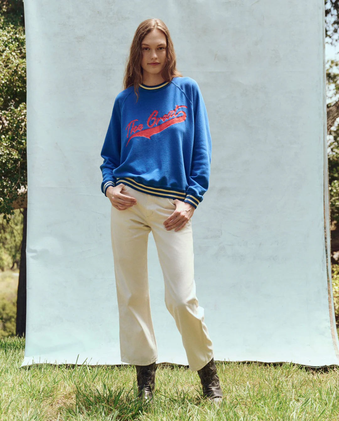 A person stands on grass, posing for a photo. They wear The Great Inc.'s Varsity College Sweatshirt with Crest Graphic and light-colored pants, paired with black boots. The background features a light-colored screen with partially visible trees.