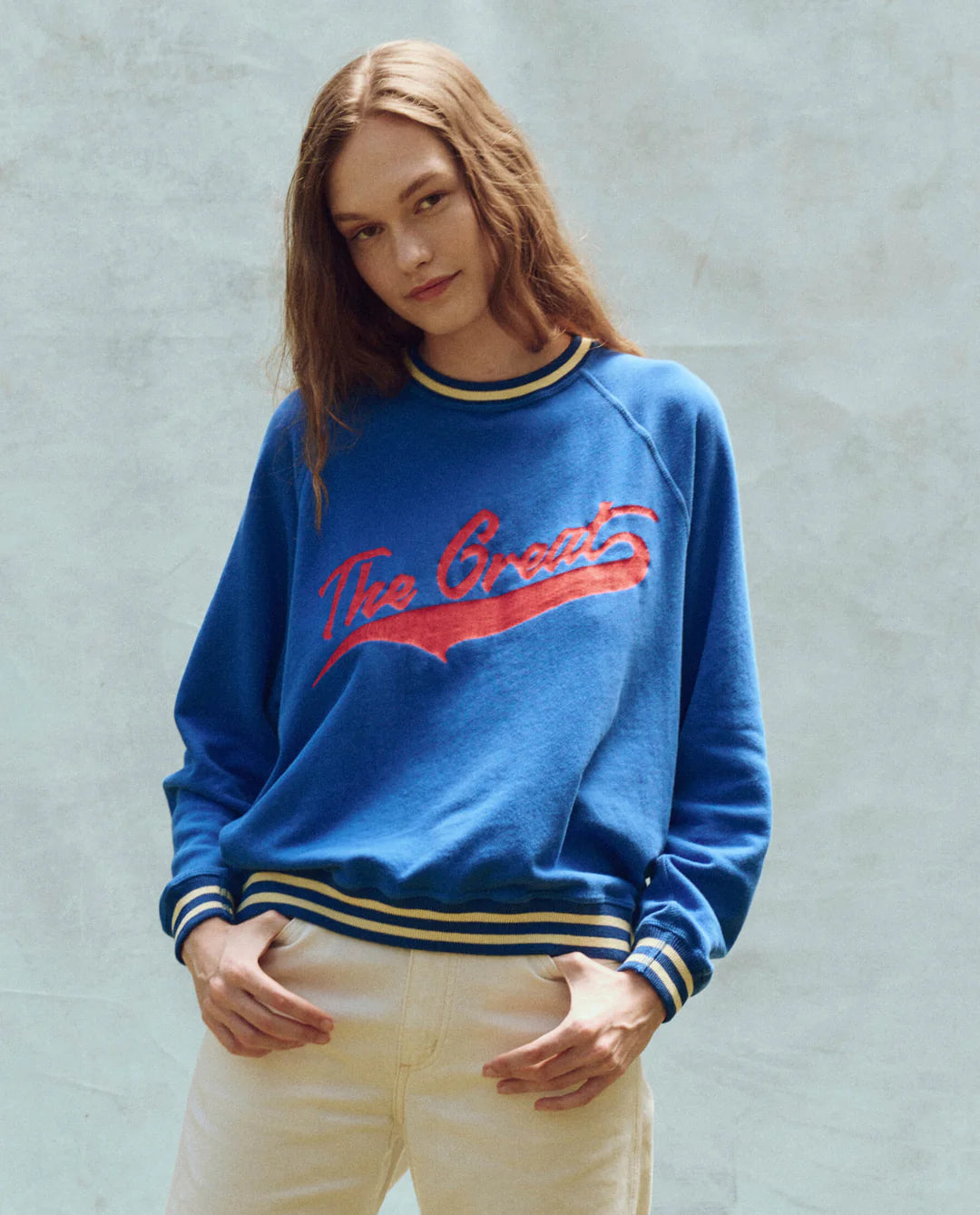 A person with long hair is wearing The Great Inc.'s The Varsity College Sweatshirt with Crest Graphic, featuring "The Greats" in red script, striped cuffs, and hem. They're also in light-colored pants against a textured off-white background.