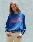 A person with long hair is wearing The Great Inc.'s The Varsity College Sweatshirt with Crest Graphic, featuring "The Greats" in red script, striped cuffs, and hem. They're also in light-colored pants against a textured off-white background.