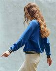 A person with long, wavy hair smiles while walking and looking down. They wear The Great Inc.'s Varsity College Sweatshirt with Crest Graphic, featuring blue tones and striped cuffs, paired with off-white pants against a light gray backdrop.