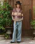 A person in a black hat, The Riding Cardigan by The Great Inc. with southwestern motifs and a belt, wide-leg jeans, and black shoes stands in front of a wooden door surrounded by potted green plants.