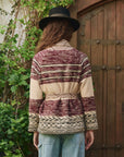 A person with long hair, wearing a black hat and The Riding Cardigan by The Great Inc.—a shawl-collar piece featuring southwestern motifs in maroon and beige—stands facing a wooden door surrounded by greenery.