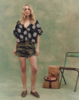 A woman stands indoors against a green mottled backdrop, wearing The Cargo Fatigue Short by The Great Inc., a navy floral blouse, and brown boat shoes. Nearby are a rustic log stool, green metal chair, and wicker bag.